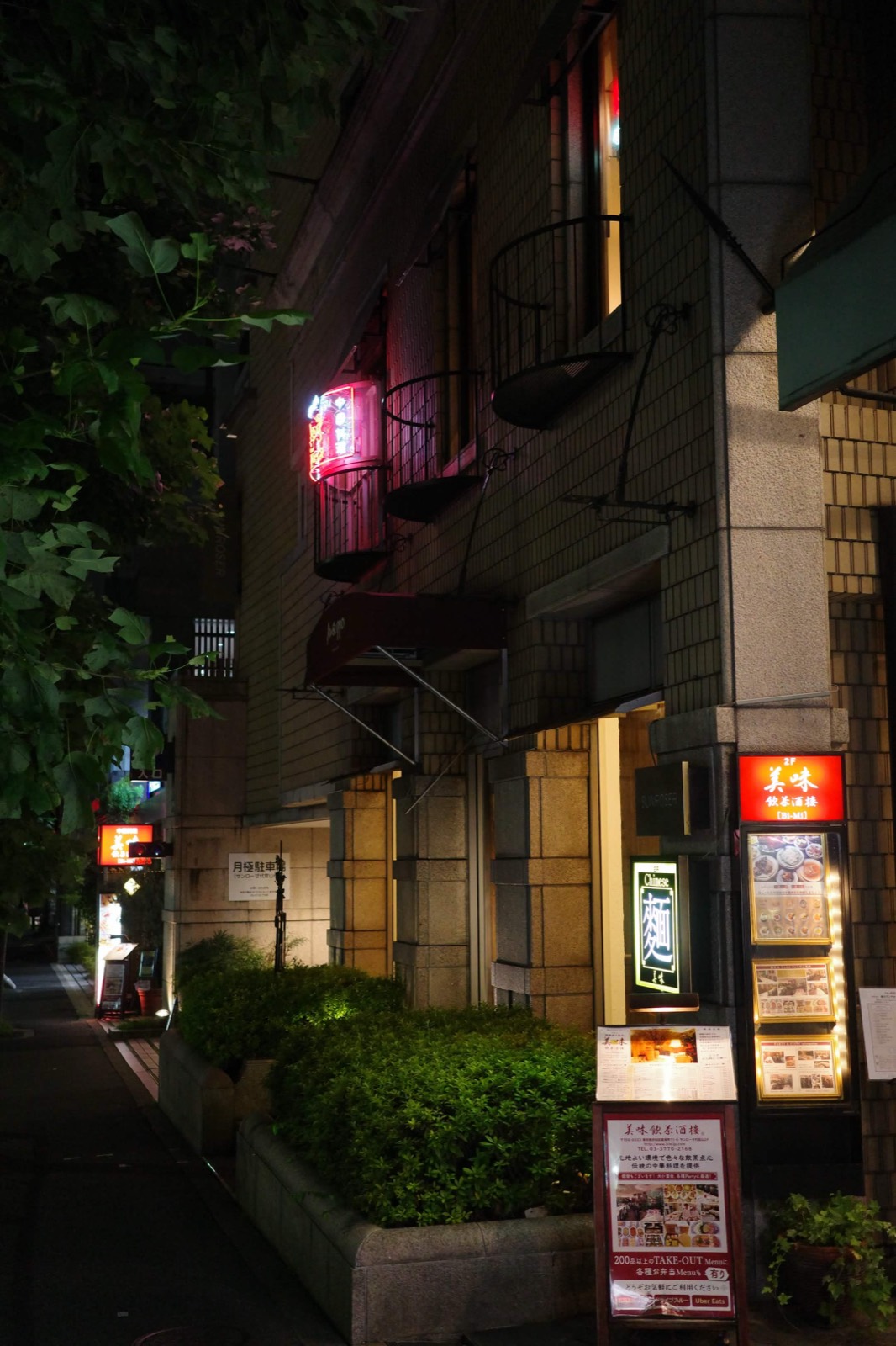 Night backstreet with a pink neon sign glowing on a tiled facade and lit menu boards below.