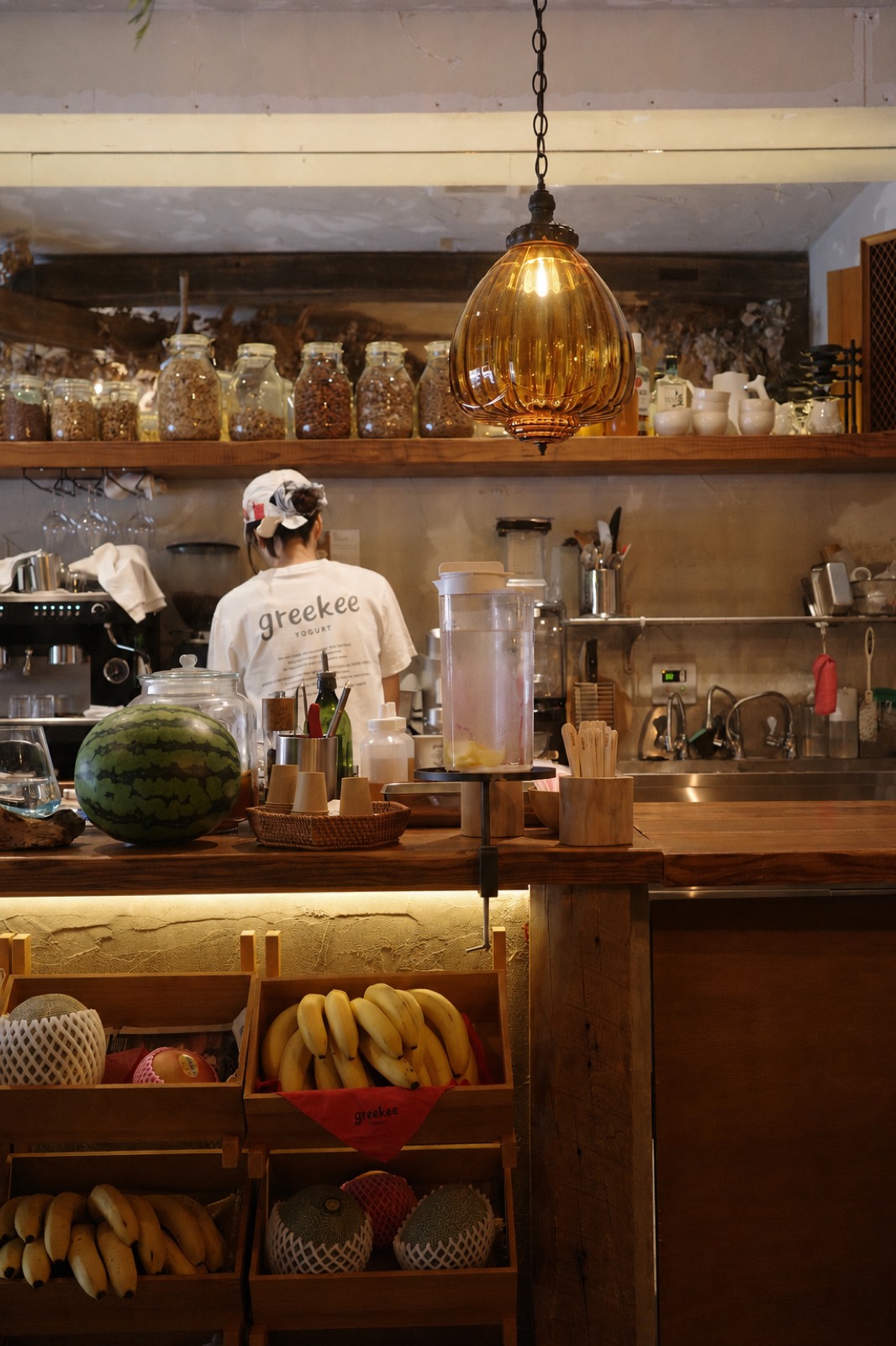 Café interior with an amber glass pendant lamp, jars along a shelf, and crates of fruit below the wooden counter.